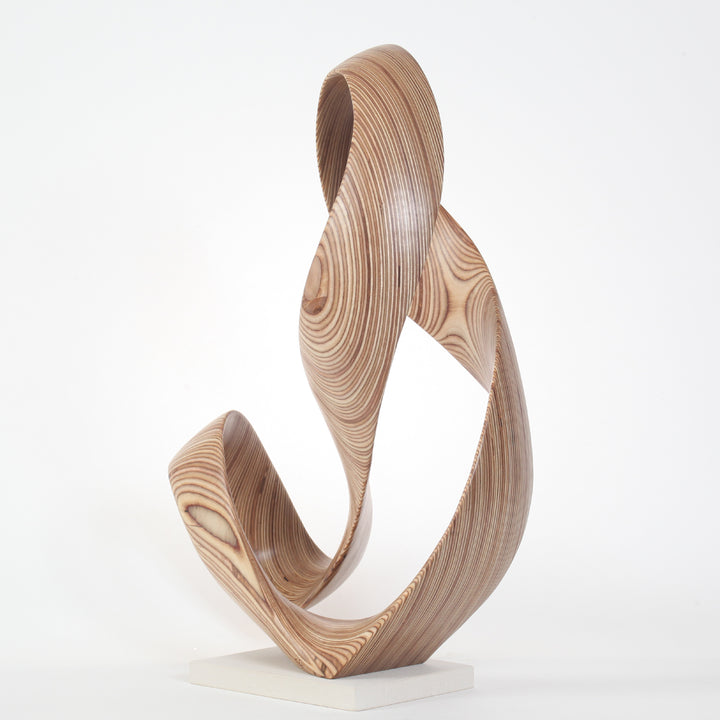 Hand-carved birch plywood sculpture by Bill Prickett, formed as a flowing Möbius-style infinity loop inspired by an ampersand, with continuous twisting curves, visible layered grain, and a balanced sculptural presence on a white base.