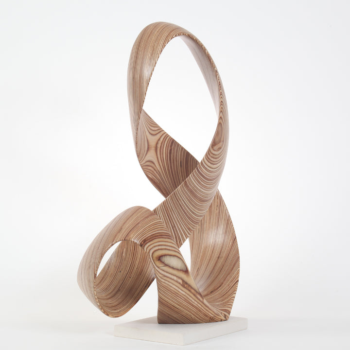 Hand-carved birch plywood sculpture by Bill Prickett, formed as a flowing Möbius-style infinity loop inspired by an ampersand, with continuous twisting curves, visible layered grain, and a balanced sculptural presence on a white base.