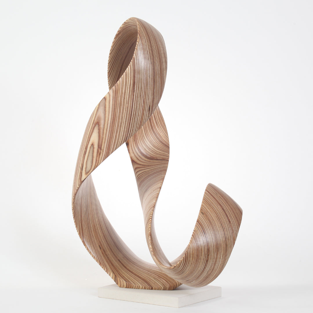 Hand-carved birch plywood sculpture by Bill Prickett, formed as a flowing Möbius-style infinity loop inspired by an ampersand, with continuous twisting curves, visible layered grain, and a balanced sculptural presence on a white base.