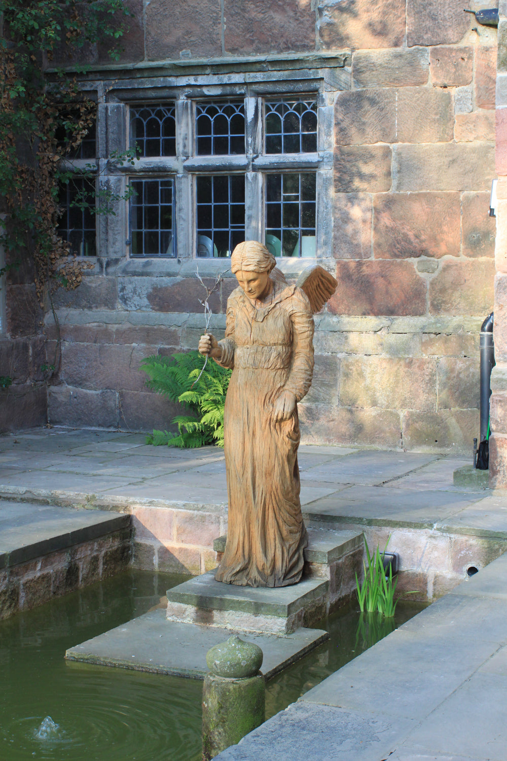 Wooden sculpture of an angel in a serene, contemplative pose, standing on a stone base near a pond. The figure wears a long robe with fine carved detailing and holds a delicate twig in one hand, with wings folded gently behind. Set against a rustic stone wall and framed by lush green ferns.