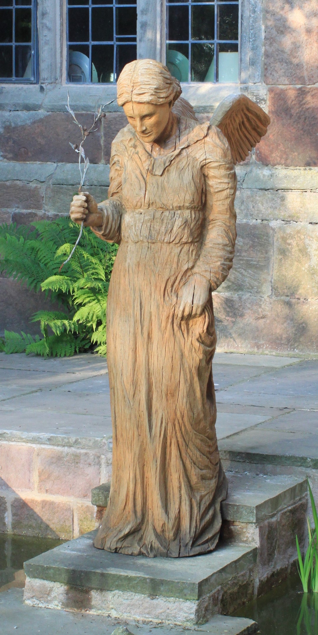 Wooden sculpture of an angel in a serene, contemplative pose, standing on a stone base near a pond. The figure wears a long robe with fine carved detailing and holds a delicate twig in one hand, with wings folded gently behind. Set against a rustic stone wall and framed by lush green ferns.