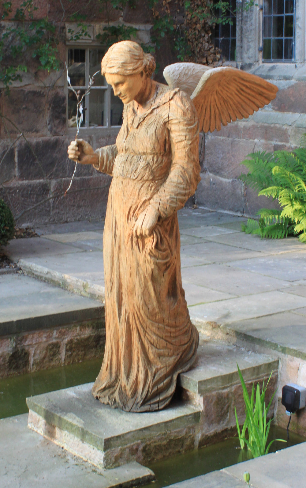 Wooden sculpture of an angel in a serene, contemplative pose, standing on a stone base near a pond. The figure wears a long robe with fine carved detailing and holds a delicate twig in one hand, with wings folded gently behind. Set against a rustic stone wall and framed by lush green ferns.