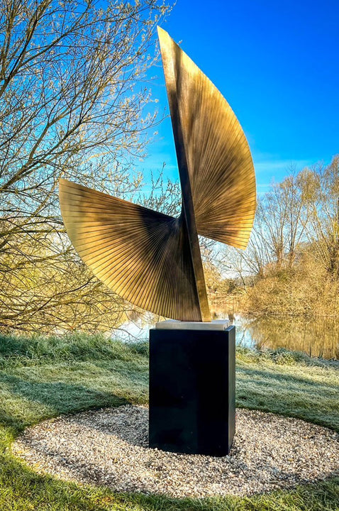 APOLLO by Thomas Joynes – a monumental outdoor sculpture fabricated in brass with an antique patina, featuring two opposing fan-like forms rising in a dynamic twist. Radiating hand-cut lines evoke celestial movement and kinetic tension. Elevated on a 70cm black steel plinth, this one-off piece stands at 2.2 metres tall, commanding presence in natural surroundings.