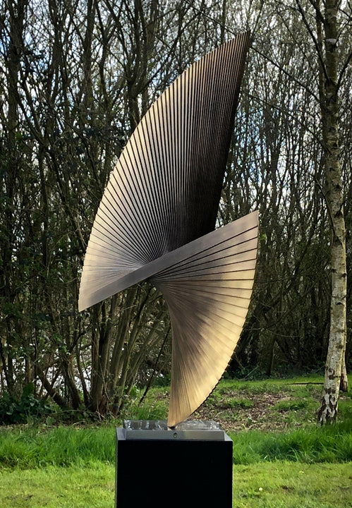APOLLO by Thomas Joynes – a monumental outdoor sculpture fabricated in brass with an antique patina, featuring two opposing fan-like forms rising in a dynamic twist. Radiating hand-cut lines evoke celestial movement and kinetic tension. Elevated on a 70cm black steel plinth, this one-off piece stands at 2.2 metres tall, commanding presence in natural surroundings.