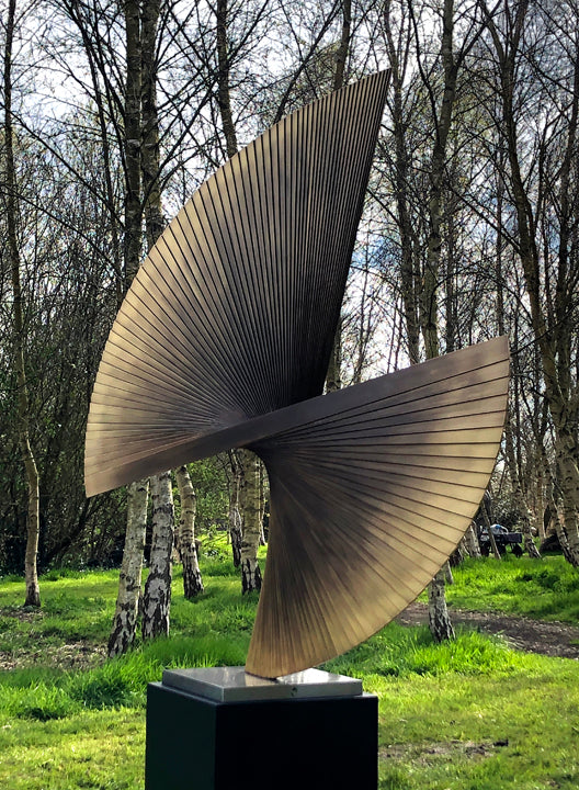 APOLLO by Thomas Joynes – a monumental outdoor sculpture fabricated in brass with an antique patina, featuring two opposing fan-like forms rising in a dynamic twist. Radiating hand-cut lines evoke celestial movement and kinetic tension. Elevated on a 70cm black steel plinth, this one-off piece stands at 2.2 metres tall, commanding presence in natural surroundings.