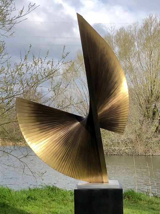APOLLO by Thomas Joynes – a monumental outdoor sculpture fabricated in brass with an antique patina, featuring two opposing fan-like forms rising in a dynamic twist. Radiating hand-cut lines evoke celestial movement and kinetic tension. Elevated on a 70cm black steel plinth, this one-off piece stands at 2.2 metres tall, commanding presence in natural surroundings.