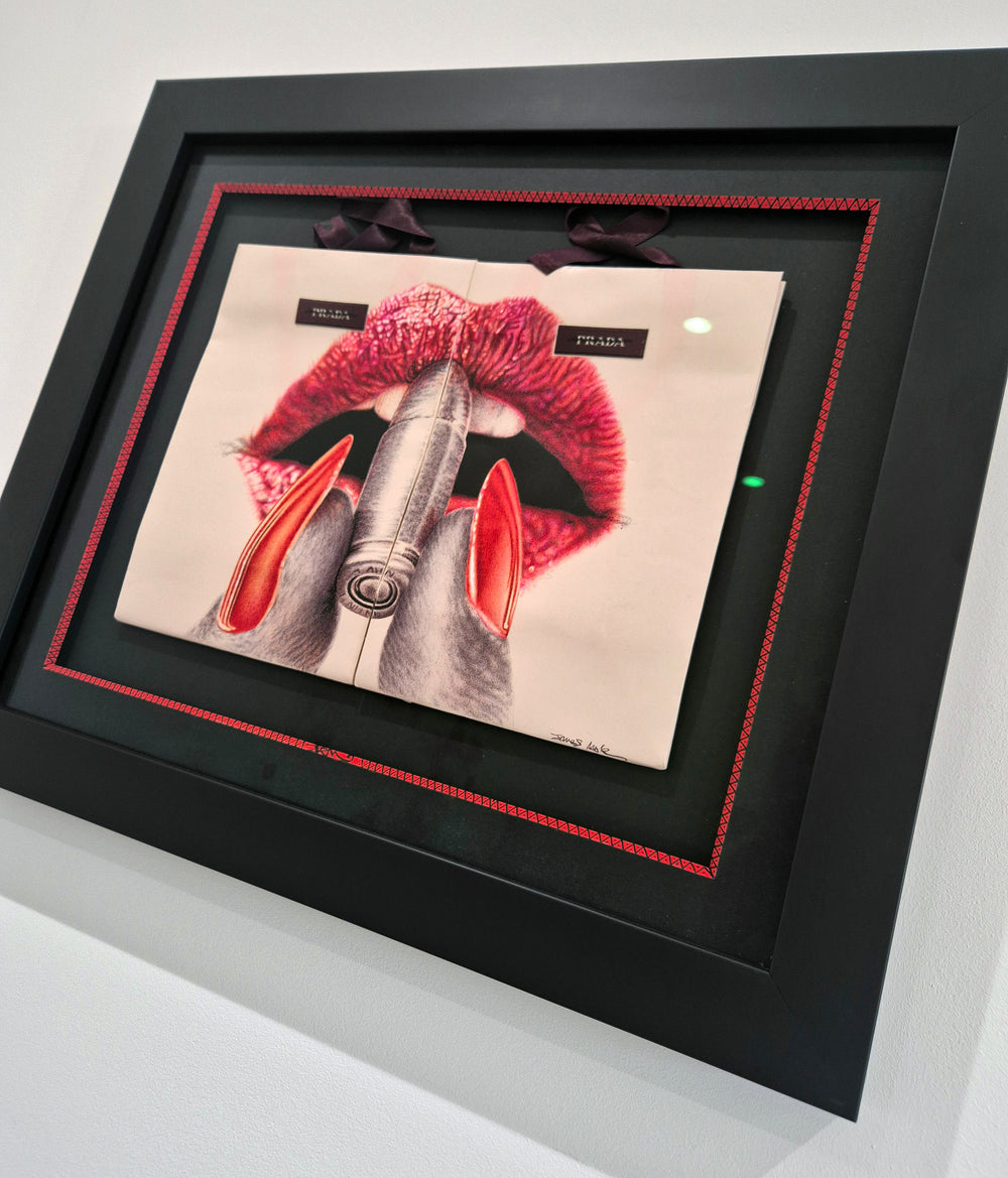 Original artwork titled “Apply With Caution” by James Mylne, featuring a provocative hyperrealistic drawing of glossy red lips applying lipstick formed from a bullet. Created using red and black ballpoint pens and acrylic marker on two Prada gift bags, the work is framed in a black box frame with red detailing and ribbon handles exposed.