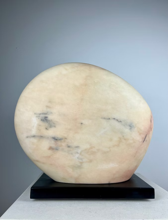 Jason Mulligan sculpture titled APRIL MOON, a one-off hand-carved abstract artwork in Rosa Aurora marble. The piece features two softly rounded, interlocking forms with smooth polished surfaces, revealing delicate tones of pale rose, peach, cream and grey-blue veining. Resting on a dark plinth, the sculpture evokes themes of lunar cycles, balance and renewal, blending timeless marble carving traditions with a contemporary minimalist aesthetic.