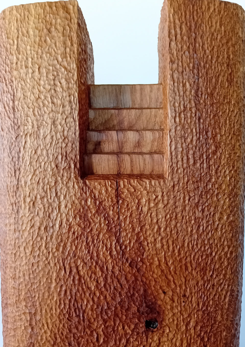 Ascent by Nicola Beattie – a tall, monolithic oak sculpture standing on a solid square oak base. Hand-carved from rich golden-brown oak with deep, textured chisel marks, the piece features a vertical architectural slot running through its length, stepped recesses at the top, and small inset hollows. The surface reveals warm amber and honey tones with darker grain lines, natural knots, and splits in the base, evoking the weathered strength of ancient standing stones and castle remnants.