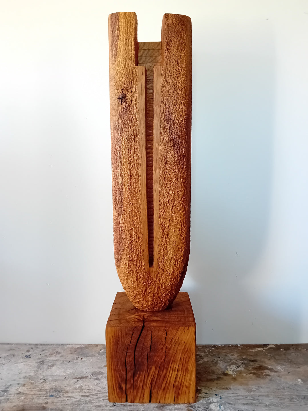 Ascent by Nicola Beattie – a tall, monolithic oak sculpture standing on a solid square oak base. Hand-carved from rich golden-brown oak with deep, textured chisel marks, the piece features a vertical architectural slot running through its length, stepped recesses at the top, and small inset hollows. The surface reveals warm amber and honey tones with darker grain lines, natural knots, and splits in the base, evoking the weathered strength of ancient standing stones and castle remnants.