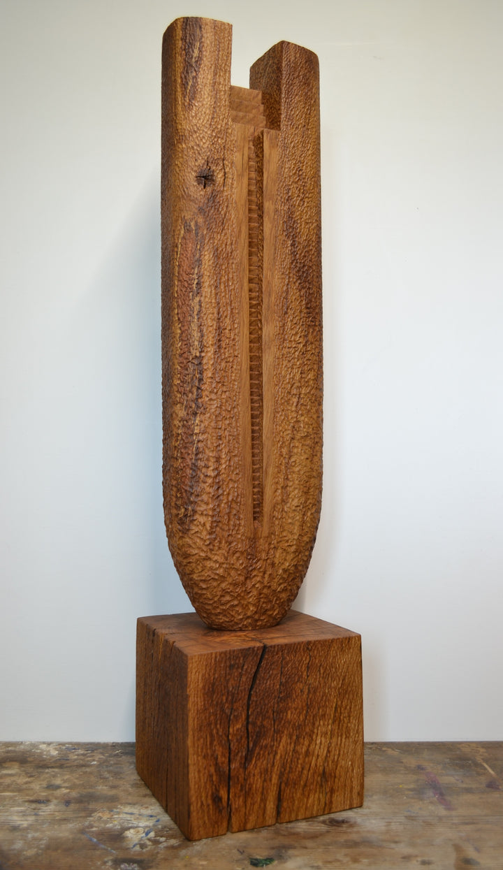 Ascent by Nicola Beattie – a tall, monolithic oak sculpture standing on a solid square oak base. Hand-carved from rich golden-brown oak with deep, textured chisel marks, the piece features a vertical architectural slot running through its length, stepped recesses at the top, and small inset hollows. The surface reveals warm amber and honey tones with darker grain lines, natural knots, and splits in the base, evoking the weathered strength of ancient standing stones and castle remnants.