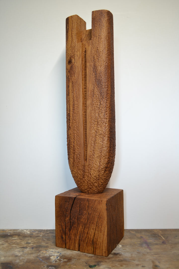 Ascent by Nicola Beattie – a tall, monolithic oak sculpture standing on a solid square oak base. Hand-carved from rich golden-brown oak with deep, textured chisel marks, the piece features a vertical architectural slot running through its length, stepped recesses at the top, and small inset hollows. The surface reveals warm amber and honey tones with darker grain lines, natural knots, and splits in the base, evoking the weathered strength of ancient standing stones and castle remnants.