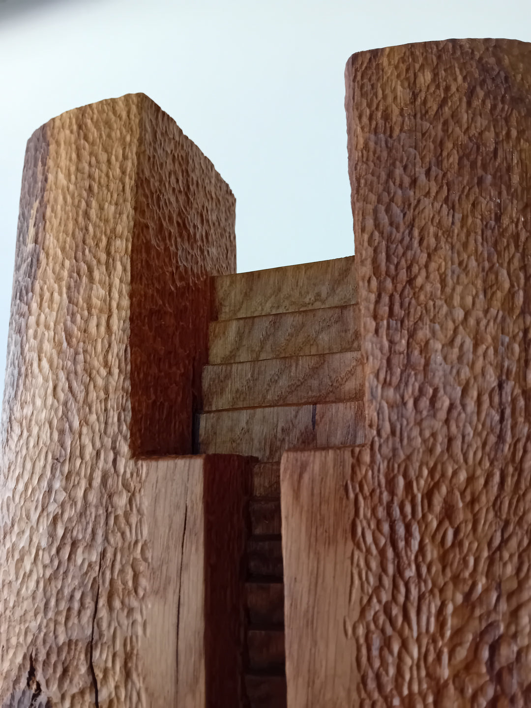 Ascent by Nicola Beattie – a tall, monolithic oak sculpture standing on a solid square oak base. Hand-carved from rich golden-brown oak with deep, textured chisel marks, the piece features a vertical architectural slot running through its length, stepped recesses at the top, and small inset hollows. The surface reveals warm amber and honey tones with darker grain lines, natural knots, and splits in the base, evoking the weathered strength of ancient standing stones and castle remnants.