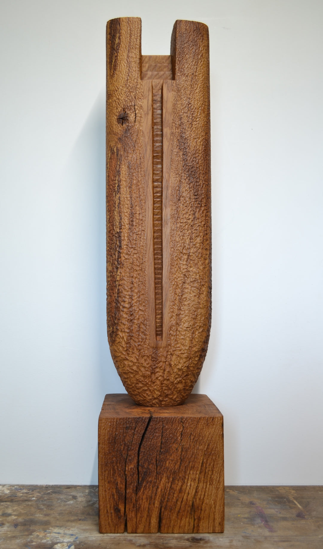 Ascent by Nicola Beattie – a tall, monolithic oak sculpture standing on a solid square oak base. Hand-carved from rich golden-brown oak with deep, textured chisel marks, the piece features a vertical architectural slot running through its length, stepped recesses at the top, and small inset hollows. The surface reveals warm amber and honey tones with darker grain lines, natural knots, and splits in the base, evoking the weathered strength of ancient standing stones and castle remnants.