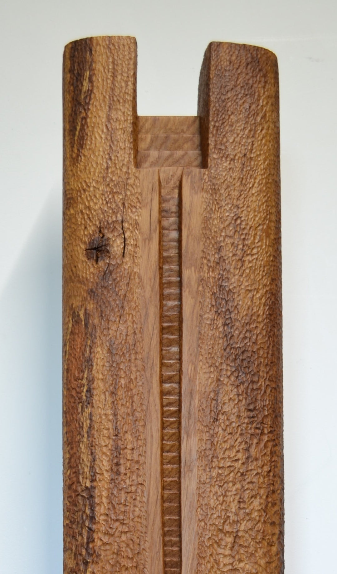Ascent by Nicola Beattie – a tall, monolithic oak sculpture standing on a solid square oak base. Hand-carved from rich golden-brown oak with deep, textured chisel marks, the piece features a vertical architectural slot running through its length, stepped recesses at the top, and small inset hollows. The surface reveals warm amber and honey tones with darker grain lines, natural knots, and splits in the base, evoking the weathered strength of ancient standing stones and castle remnants.
