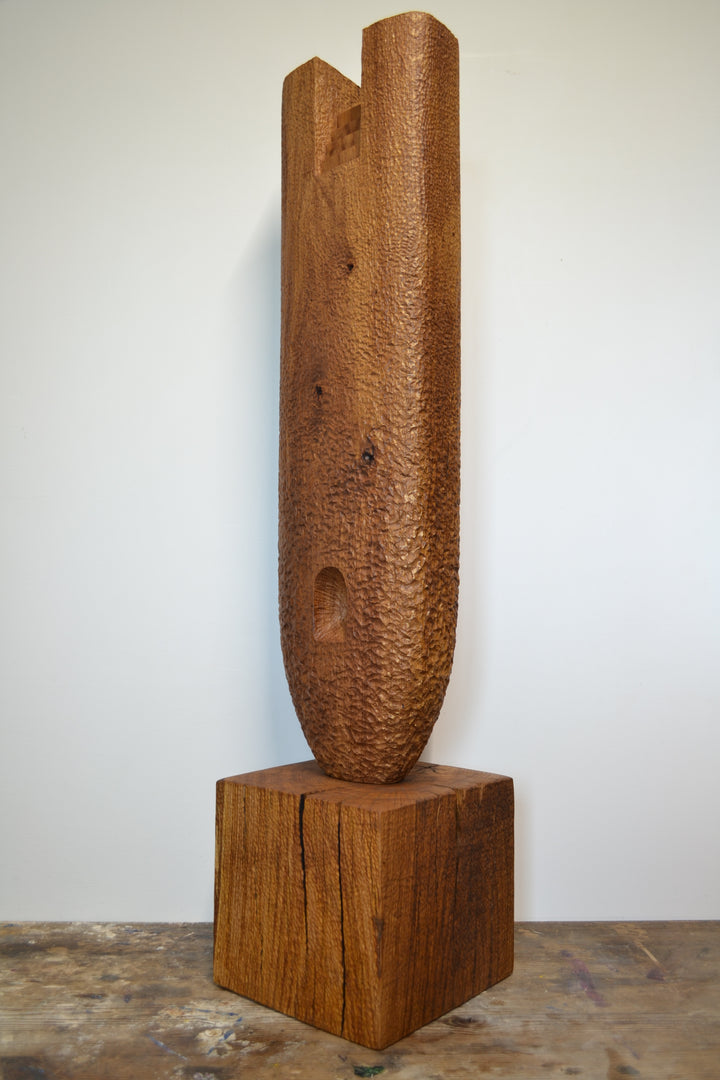 Ascent by Nicola Beattie – a tall, monolithic oak sculpture standing on a solid square oak base. Hand-carved from rich golden-brown oak with deep, textured chisel marks, the piece features a vertical architectural slot running through its length, stepped recesses at the top, and small inset hollows. The surface reveals warm amber and honey tones with darker grain lines, natural knots, and splits in the base, evoking the weathered strength of ancient standing stones and castle remnants.