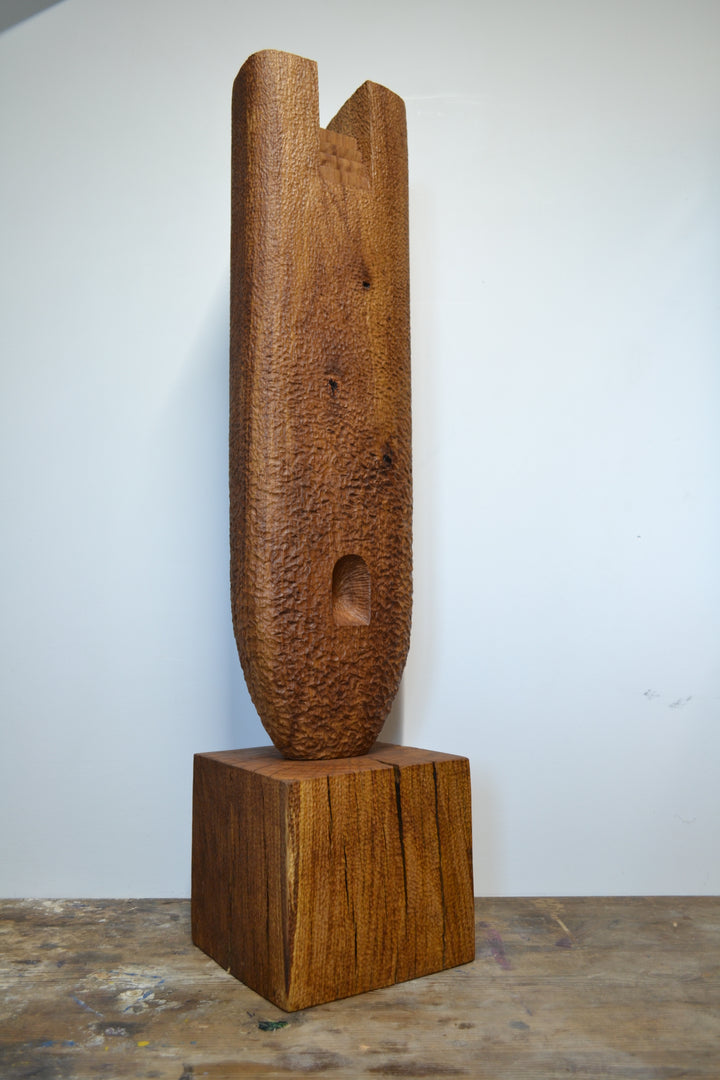 Ascent by Nicola Beattie – a tall, monolithic oak sculpture standing on a solid square oak base. Hand-carved from rich golden-brown oak with deep, textured chisel marks, the piece features a vertical architectural slot running through its length, stepped recesses at the top, and small inset hollows. The surface reveals warm amber and honey tones with darker grain lines, natural knots, and splits in the base, evoking the weathered strength of ancient standing stones and castle remnants.