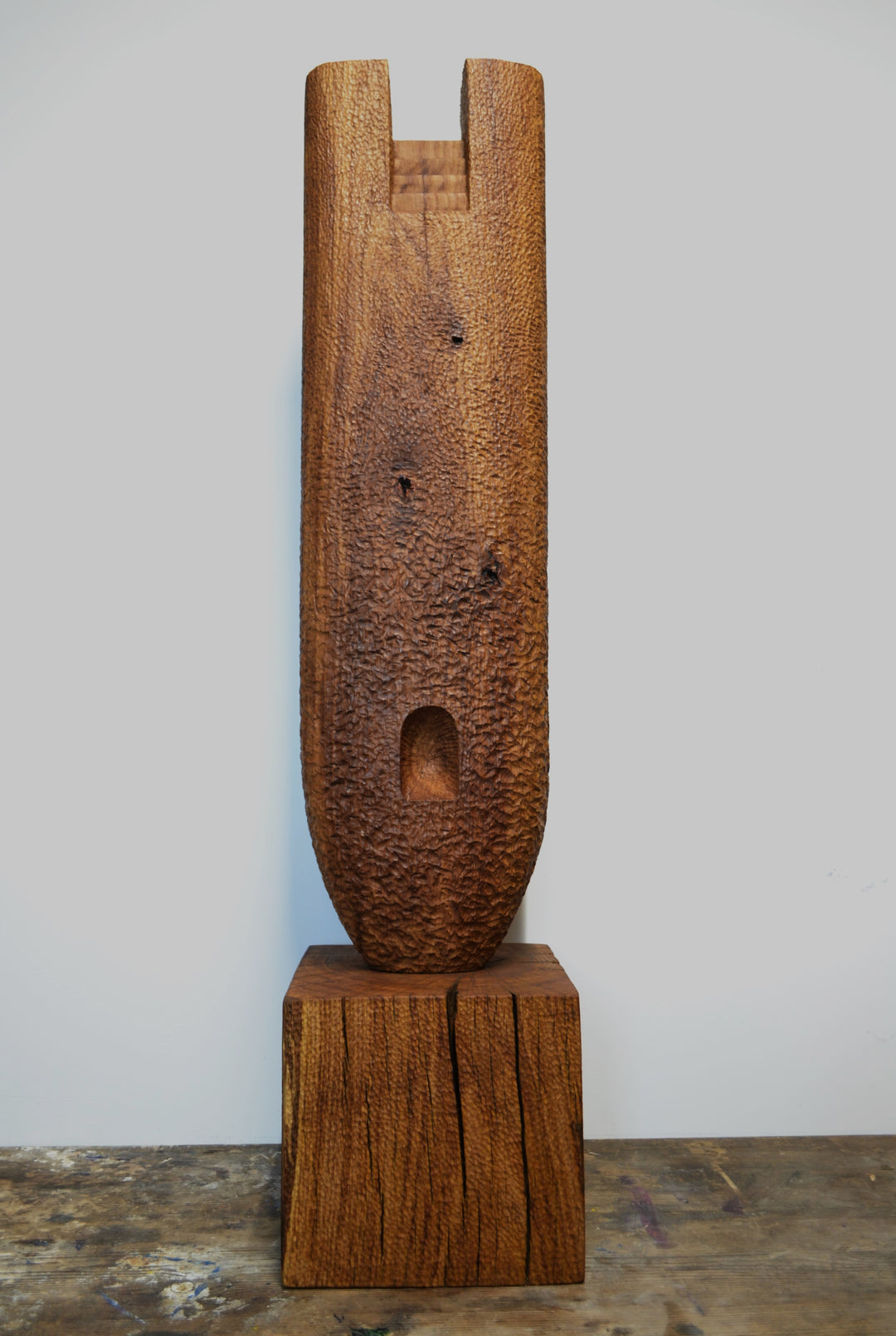 Ascent by Nicola Beattie – a tall, monolithic oak sculpture standing on a solid square oak base. Hand-carved from rich golden-brown oak with deep, textured chisel marks, the piece features a vertical architectural slot running through its length, stepped recesses at the top, and small inset hollows. The surface reveals warm amber and honey tones with darker grain lines, natural knots, and splits in the base, evoking the weathered strength of ancient standing stones and castle remnants.