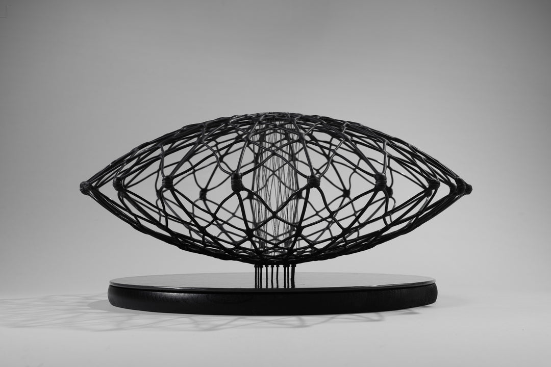Aulisca Series 2 by Tracy Nicholls — a unique kiln-formed glass sculpture with thread and wood. The piece features two symmetrical domed halves of black glass latticework, fused and slumped into a precise geometric form. Delicate strands of thread stitch the halves together at twenty points, creating a central column that adds both visual tension and structural detail. The sandblasted matte surface contrasts with the subtle inner sheen, casting intricate shadows that shift with the light