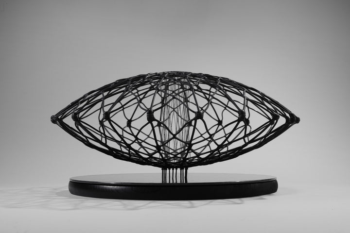 Aulisca Series 2 by Tracy Nicholls — a unique kiln-formed glass sculpture with thread and wood. The piece features two symmetrical domed halves of black glass latticework, fused and slumped into a precise geometric form. Delicate strands of thread stitch the halves together at twenty points, creating a central column that adds both visual tension and structural detail. The sandblasted matte surface contrasts with the subtle inner sheen, casting intricate shadows that shift with the light