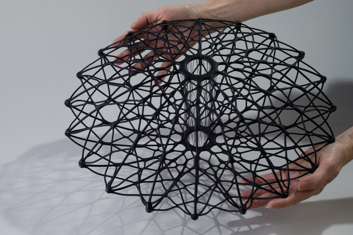 Aulisca Series 2 by Tracy Nicholls — a unique kiln-formed glass sculpture with thread and wood. The piece features two symmetrical domed halves of black glass latticework, fused and slumped into a precise geometric form. Delicate strands of thread stitch the halves together at twenty points, creating a central column that adds both visual tension and structural detail. The sandblasted matte surface contrasts with the subtle inner sheen, casting intricate shadows that shift with the light