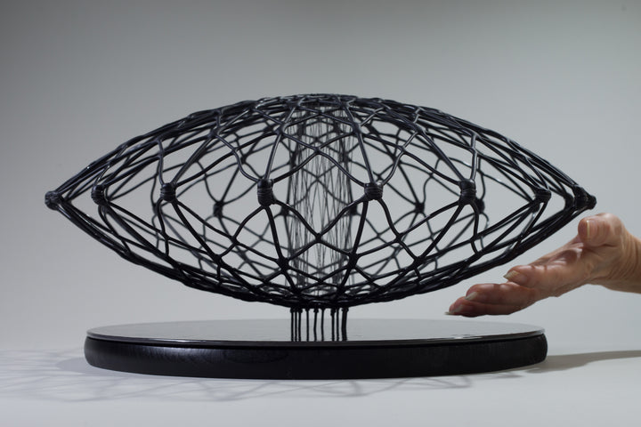 Aulisca Series 2 by Tracy Nicholls — a unique kiln-formed glass sculpture with thread and wood. The piece features two symmetrical domed halves of black glass latticework, fused and slumped into a precise geometric form. Delicate strands of thread stitch the halves together at twenty points, creating a central column that adds both visual tension and structural detail. The sandblasted matte surface contrasts with the subtle inner sheen, casting intricate shadows that shift with the light