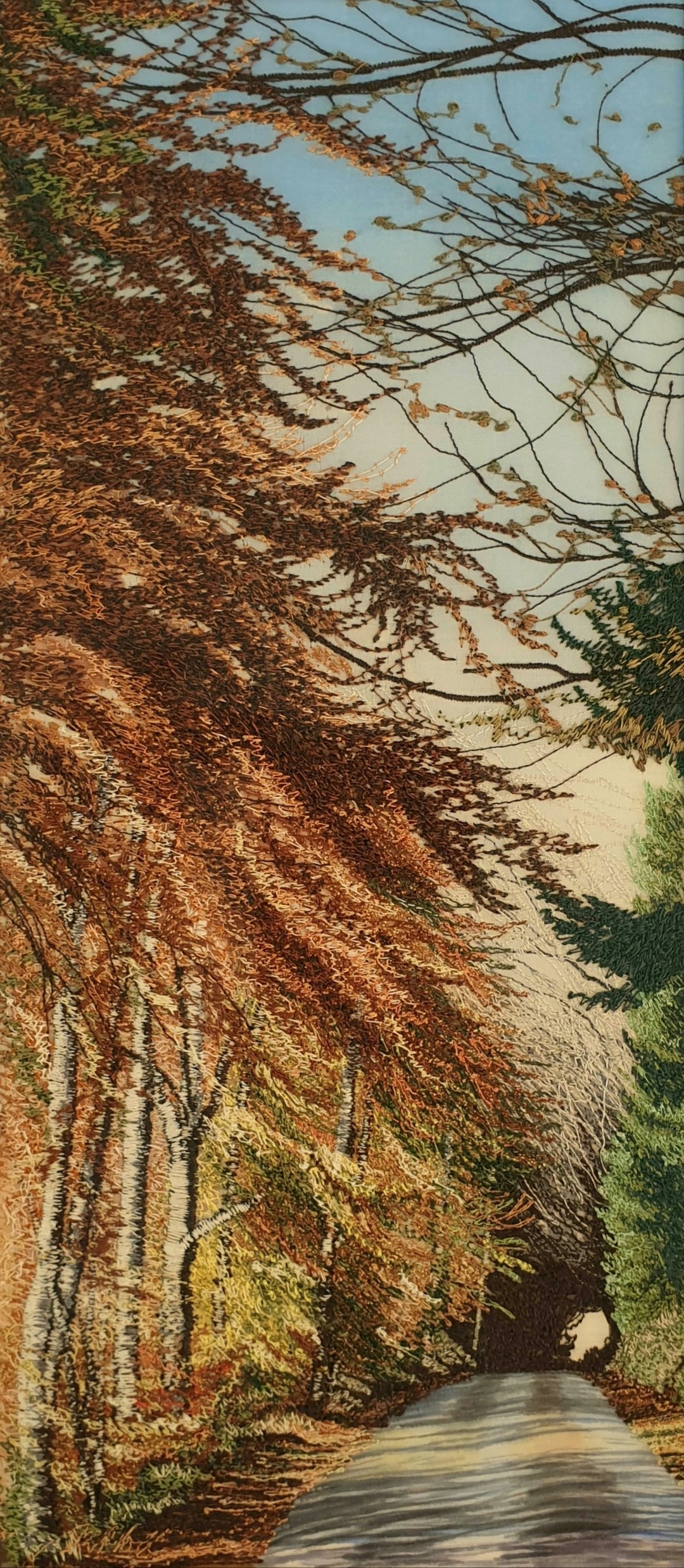 Tall embroidered artwork of a quiet road lined with autumnal trees in golden brown and orange thread, under a pale blue sky.