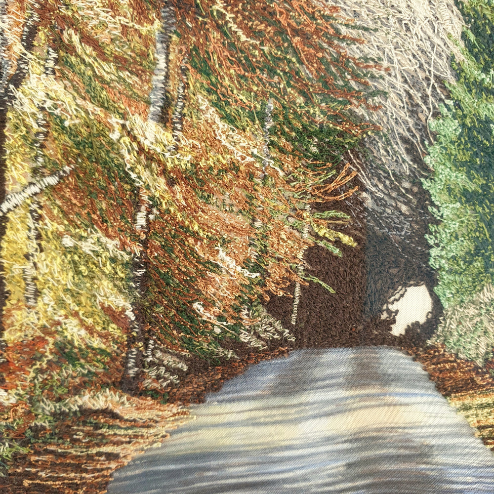 Tall embroidered artwork of a quiet road lined with autumnal trees in golden brown and orange thread, under a pale blue sky.