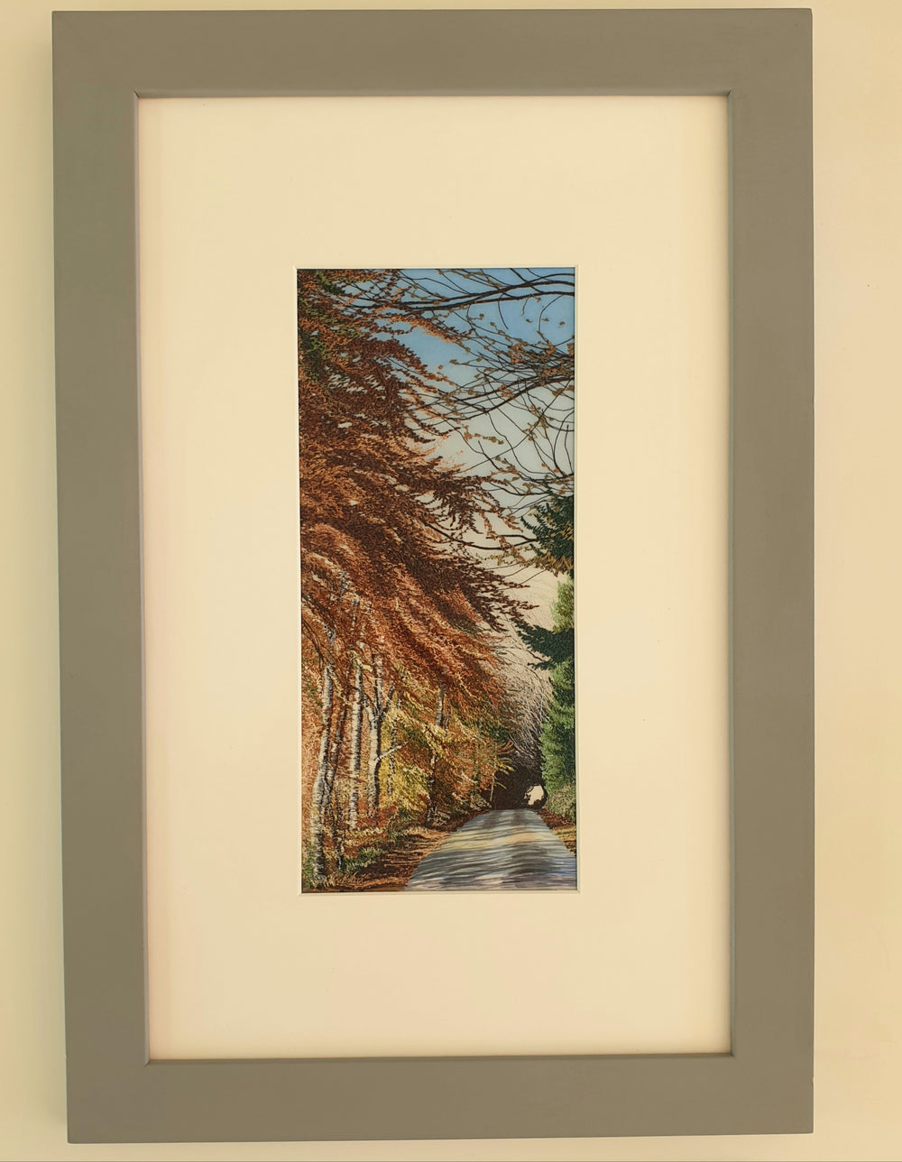Tall embroidered artwork of a quiet road lined with autumnal trees in golden brown and orange thread, under a pale blue sky.