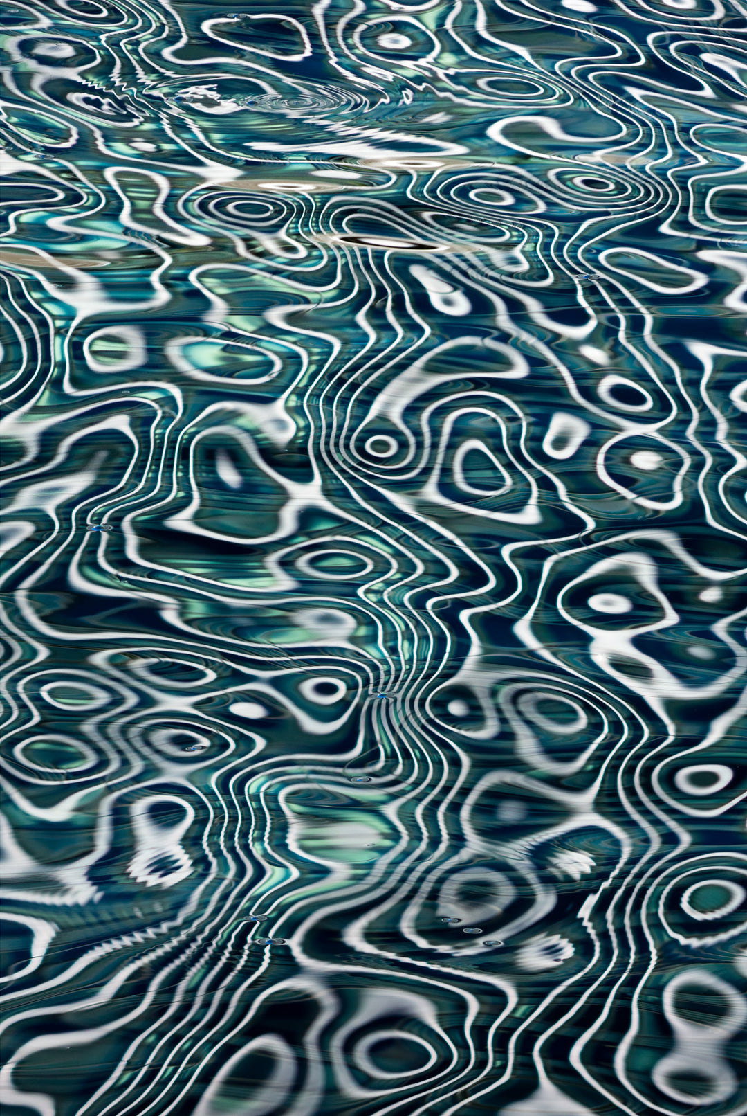 Abstract water reflection photograph with intricate ripple and wave patterns in shades of teal, blue, and white, creating a hypnotic and fluid topographic effect.