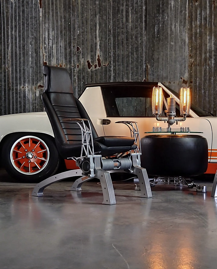 Pair of bespoke chairs handcrafted from original Porsche 914 seats and precision-engineered components, including billet aluminium legs, a modified 914 torsion tube, and hand-shaped walnut armrests, photographed in an industrial garage setting with a classic Porsche in the background.