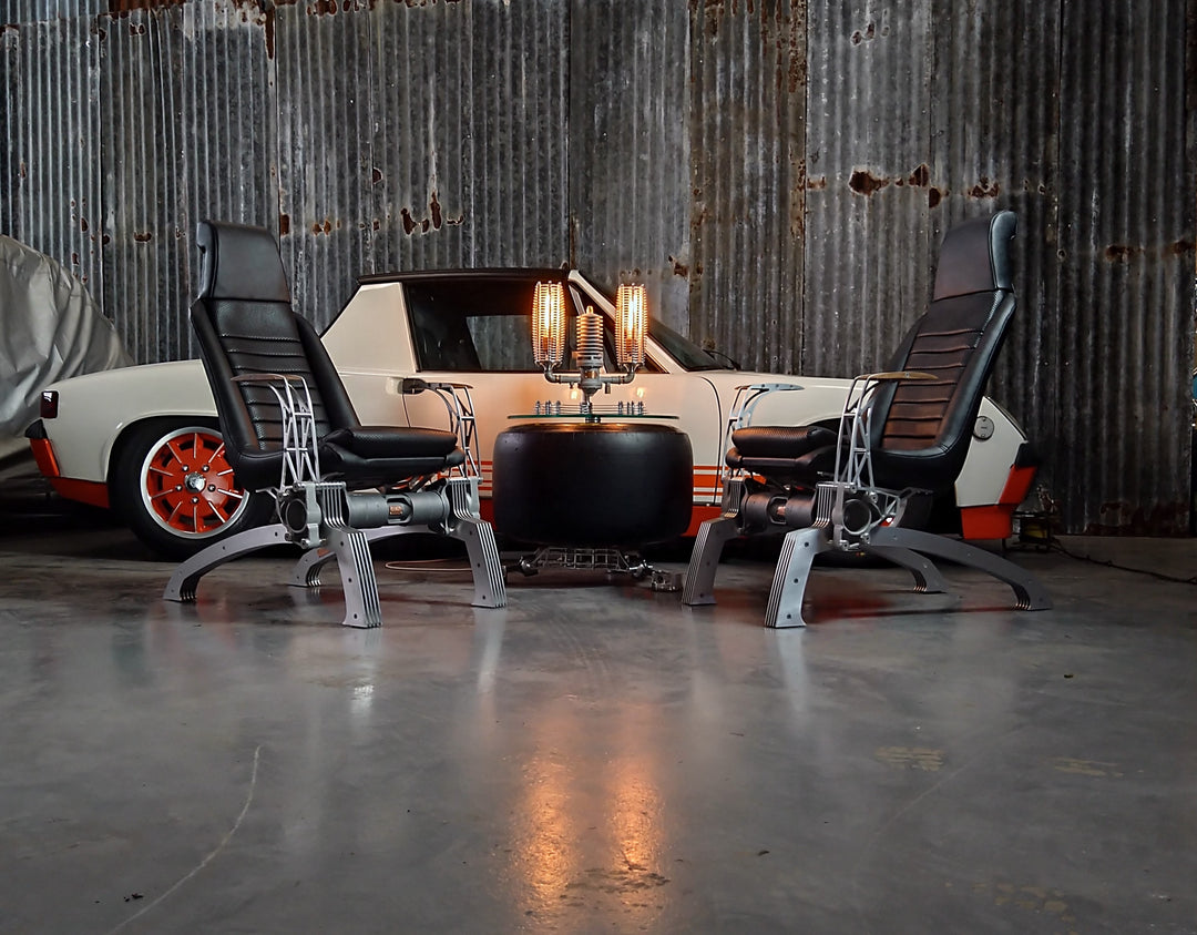 Pair of bespoke chairs handcrafted from original Porsche 914 seats and precision-engineered components, including billet aluminium legs, a modified 914 torsion tube, and hand-shaped walnut armrests, photographed in an industrial garage setting with a classic Porsche in the background.
