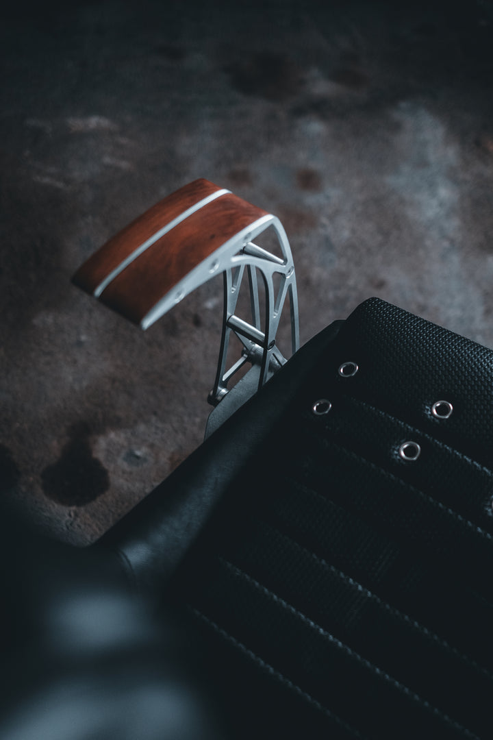 Pair of bespoke chairs handcrafted from original Porsche 914 seats and precision-engineered components, including billet aluminium legs, a modified 914 torsion tube, and hand-shaped walnut armrests, photographed in an industrial garage setting with a classic Porsche in the background.