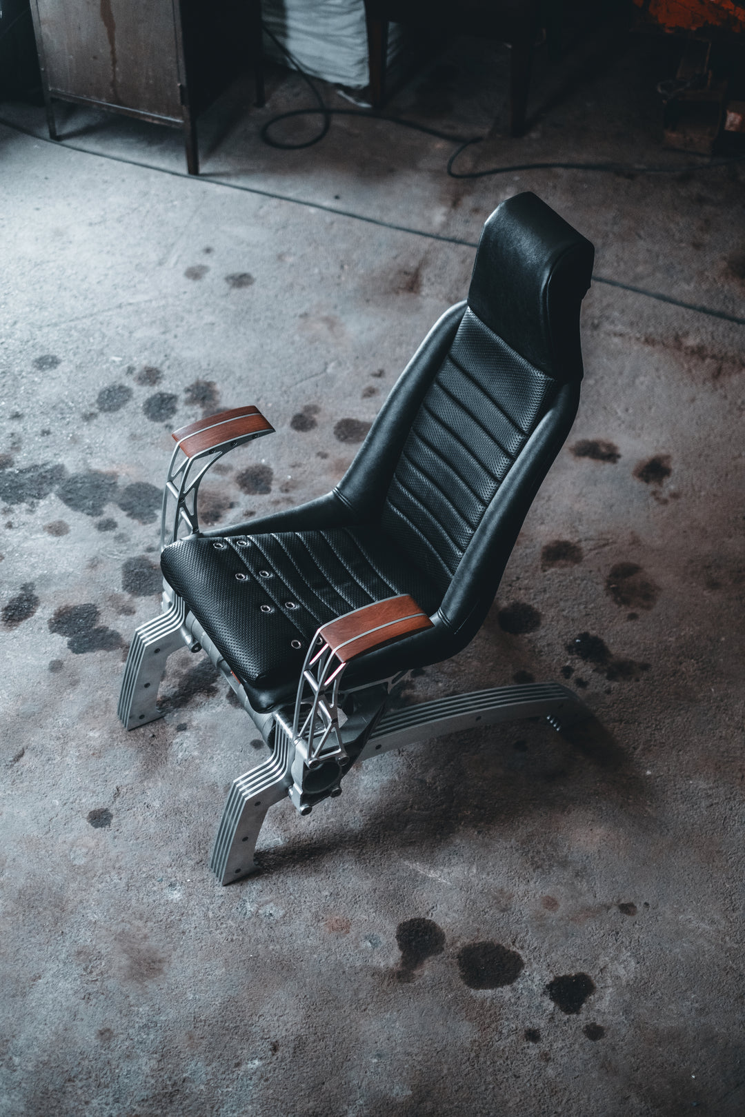 Pair of bespoke chairs handcrafted from original Porsche 914 seats and precision-engineered components, including billet aluminium legs, a modified 914 torsion tube, and hand-shaped walnut armrests, photographed in an industrial garage setting with a classic Porsche in the background.