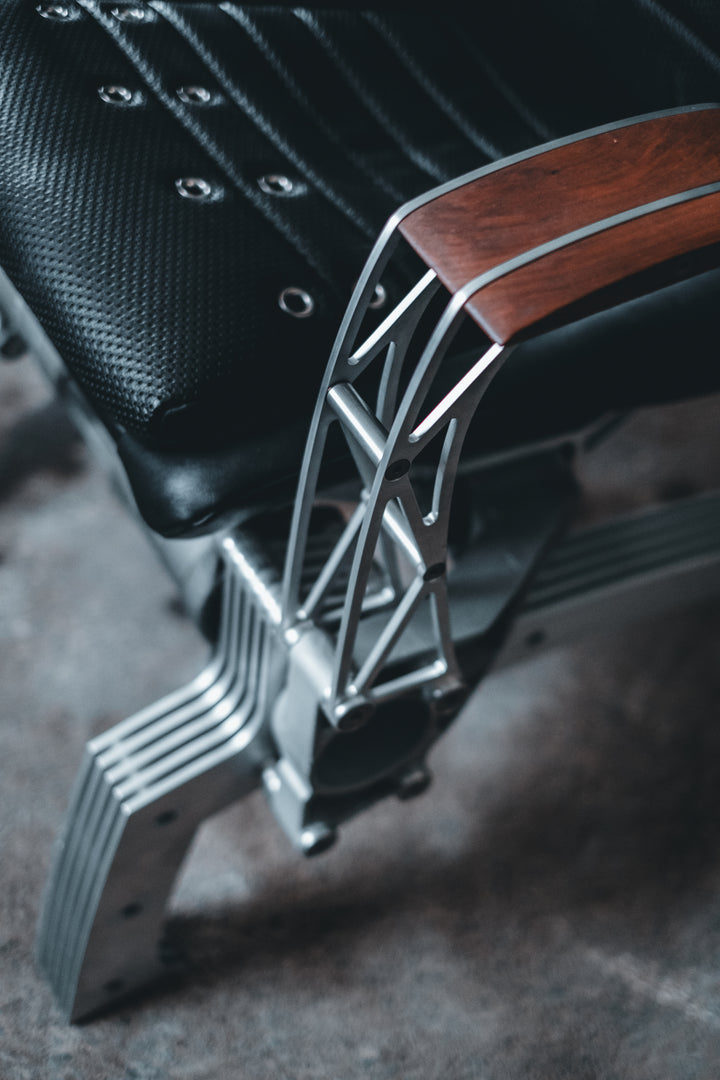 Pair of bespoke chairs handcrafted from original Porsche 914 seats and precision-engineered components, including billet aluminium legs, a modified 914 torsion tube, and hand-shaped walnut armrests, photographed in an industrial garage setting with a classic Porsche in the background.