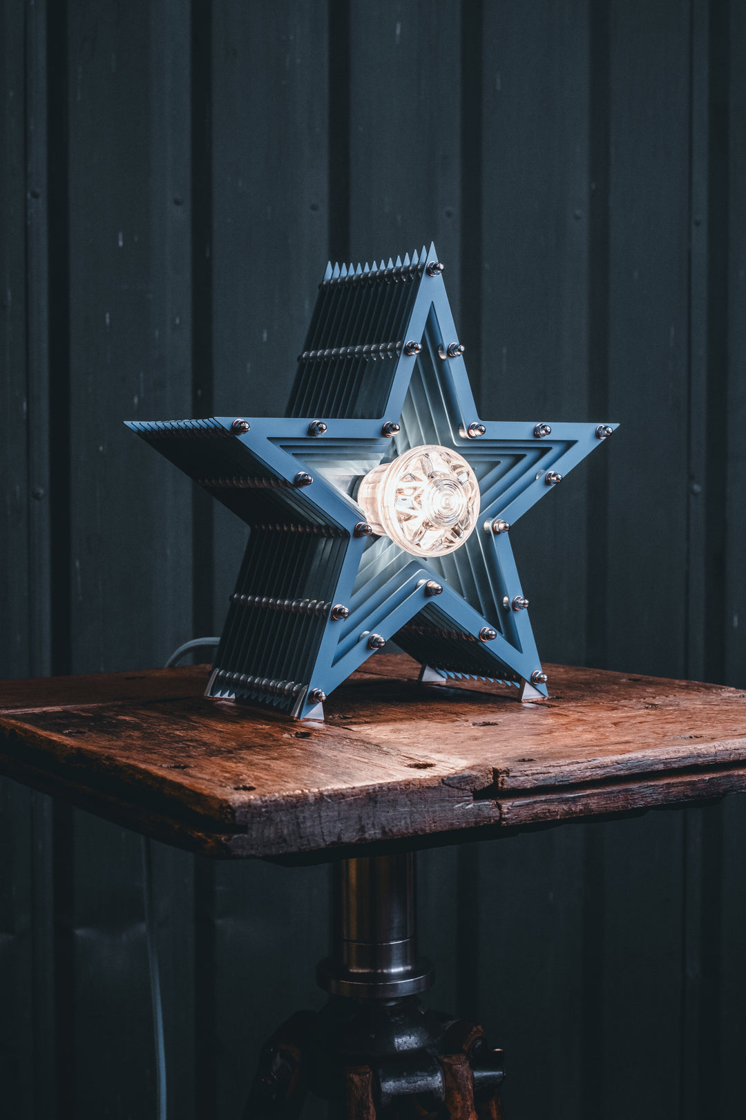 Handcrafted star-shaped lamp in deep blue metal with layered fins and exposed bulb, resting on a rustic wooden surface.