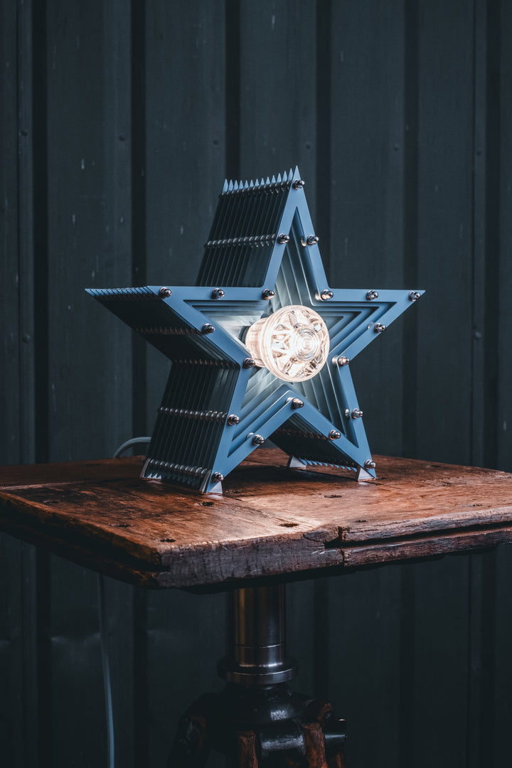 Handcrafted star-shaped lamp in deep blue metal with layered fins and exposed bulb, resting on a rustic wooden surface.