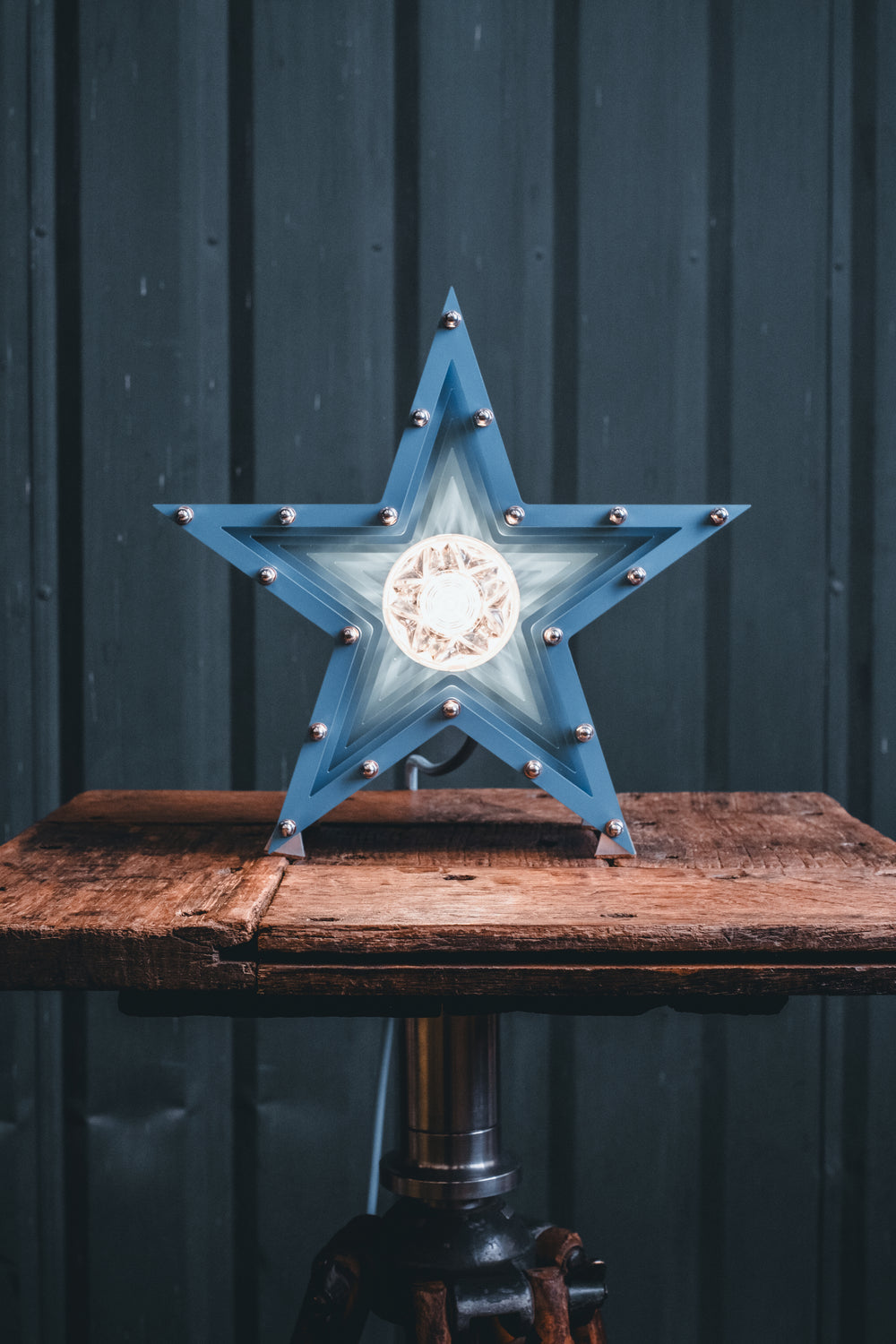 Handcrafted star-shaped lamp in deep blue metal with layered fins and exposed bulb, resting on a rustic wooden surface.
