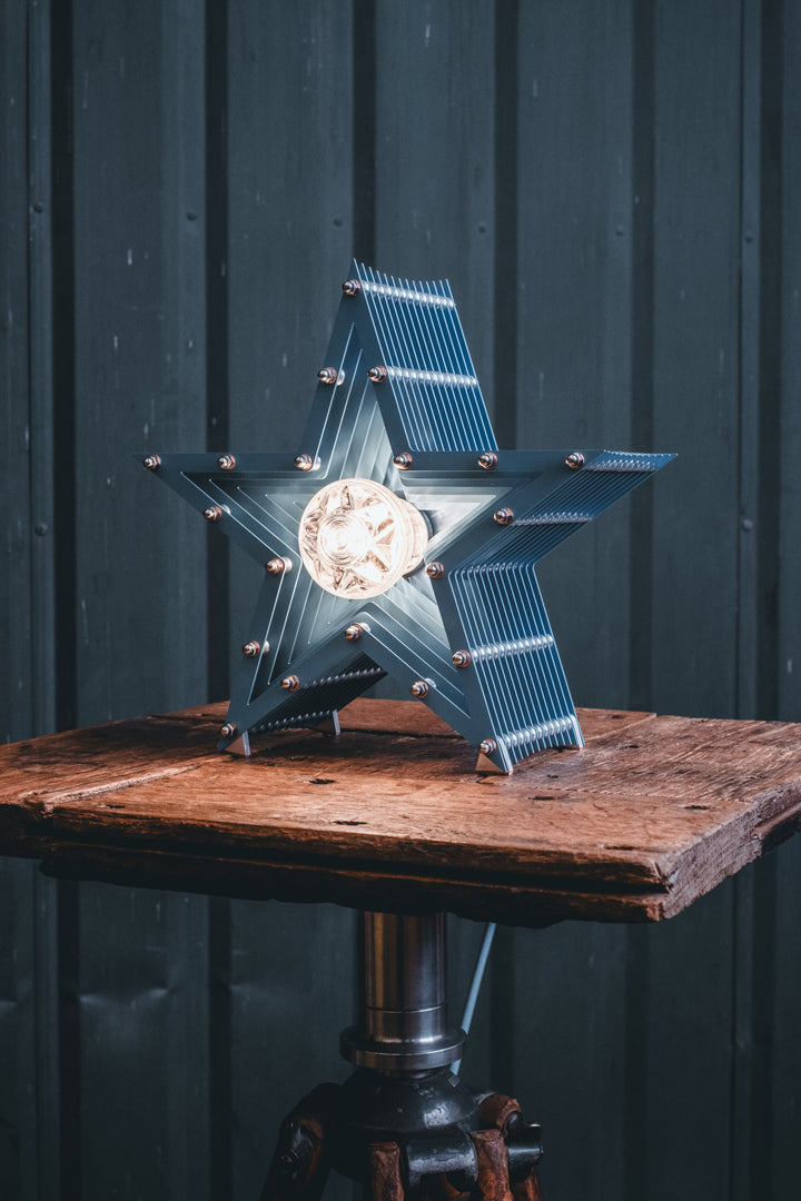 Handcrafted star-shaped lamp in deep blue metal with layered fins and exposed bulb, resting on a rustic wooden surface.