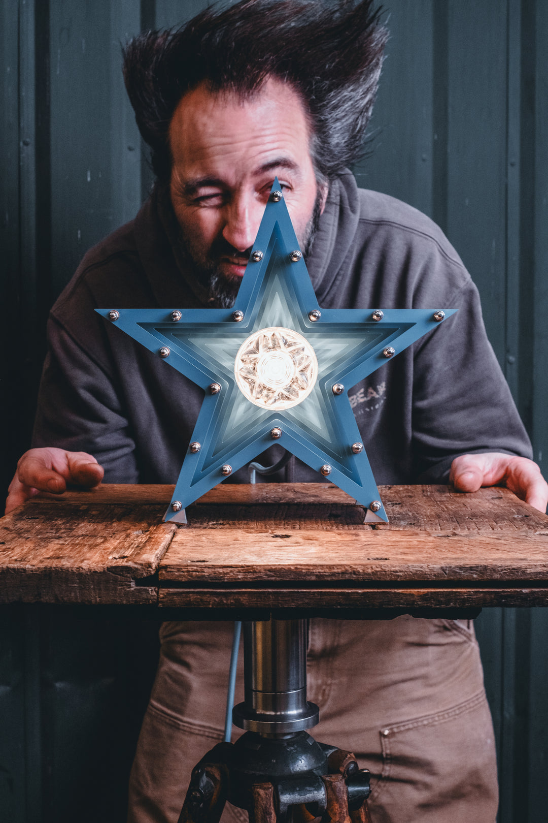 Handcrafted star-shaped lamp in deep blue metal with layered fins and exposed bulb, resting on a rustic wooden surface.