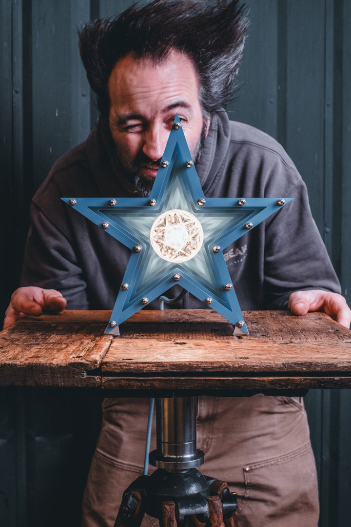 Handcrafted star-shaped lamp in deep blue metal with layered fins and exposed bulb, resting on a rustic wooden surface.