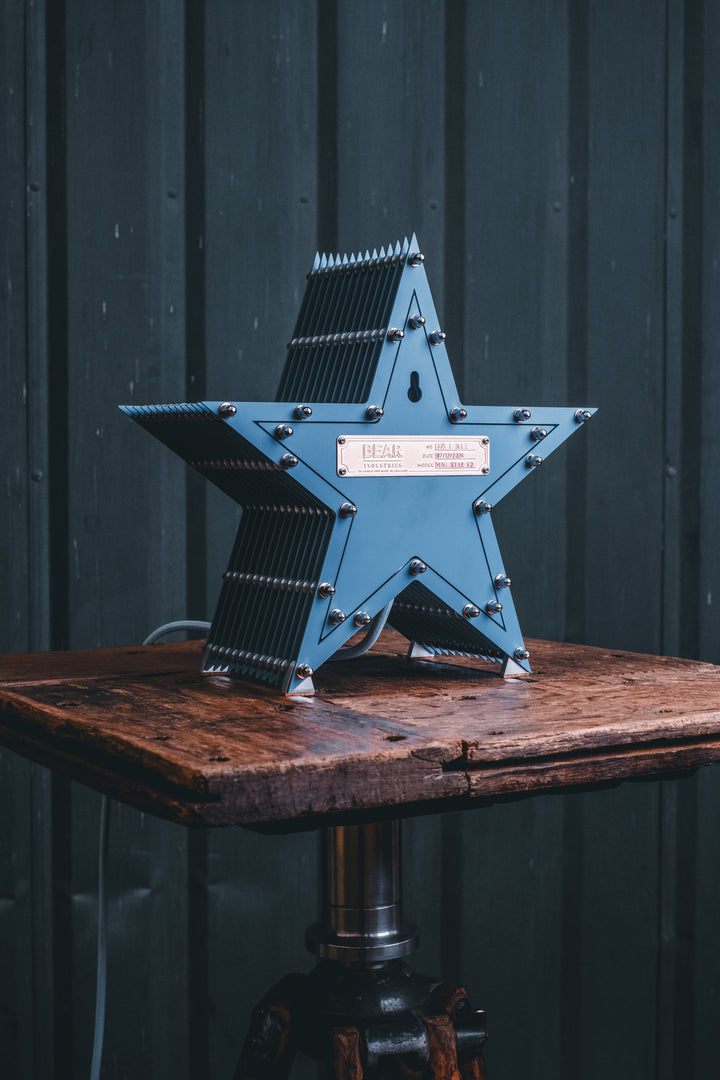 Handcrafted star-shaped lamp in deep blue metal with layered fins and exposed bulb, resting on a rustic wooden surface.