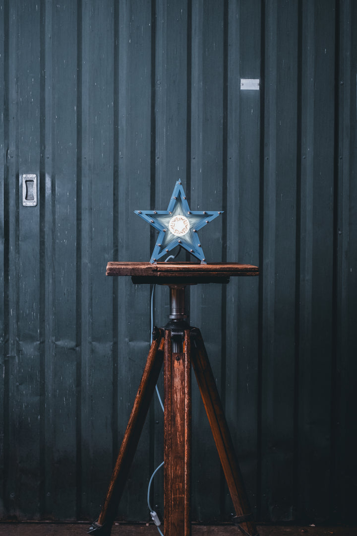 Handcrafted star-shaped lamp in deep blue metal with layered fins and exposed bulb, resting on a rustic wooden surface.