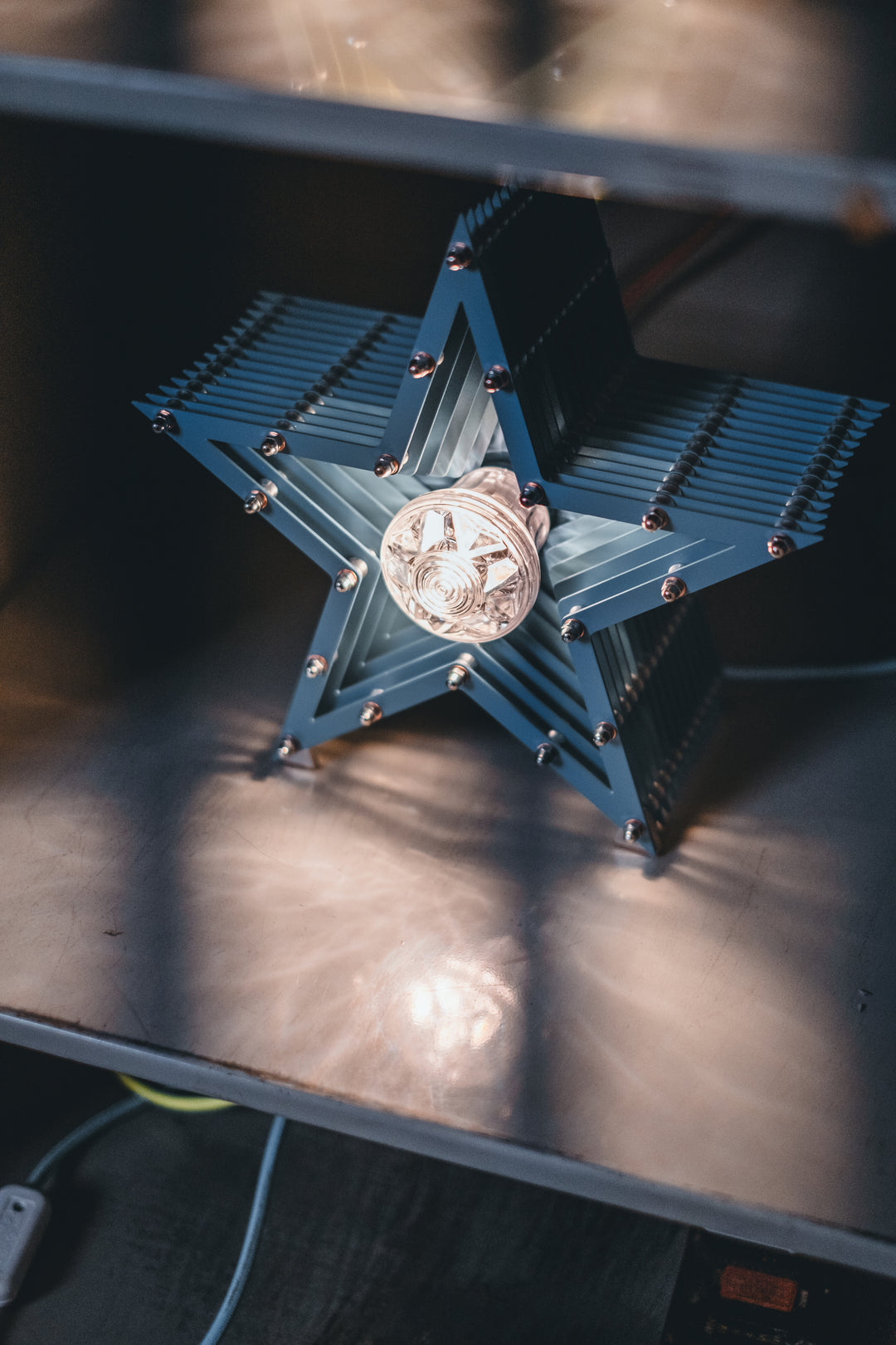 Handcrafted star-shaped lamp in deep blue metal with layered fins and exposed bulb, resting on a rustic wooden surface.