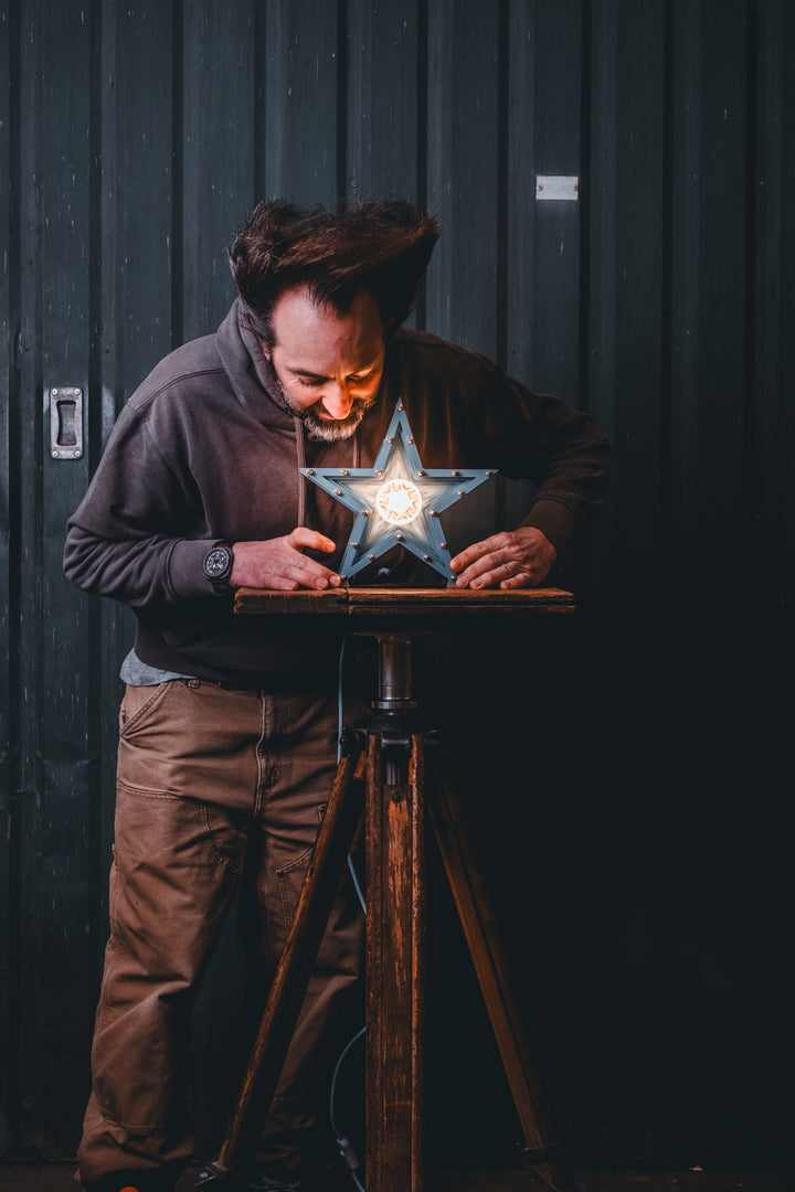 Handcrafted star-shaped lamp in deep blue metal with layered fins and exposed bulb, resting on a rustic wooden surface.
