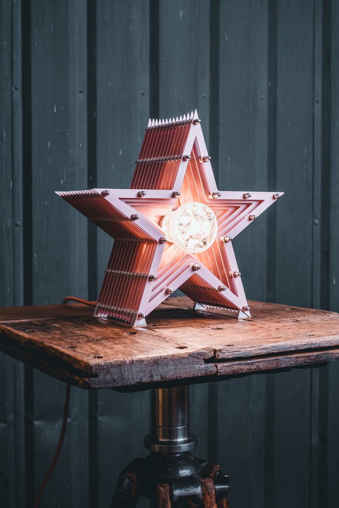 Handcrafted pink star-shaped lamp with layered metal fins and exposed central bulb, sitting on a rustic wooden table.