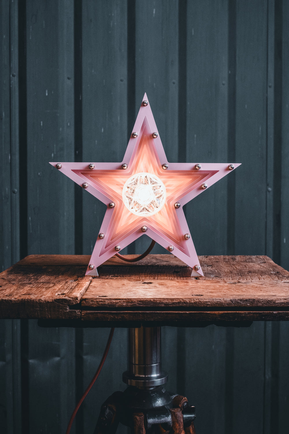 Handcrafted pink star-shaped lamp with layered metal fins and exposed central bulb, sitting on a rustic wooden table.