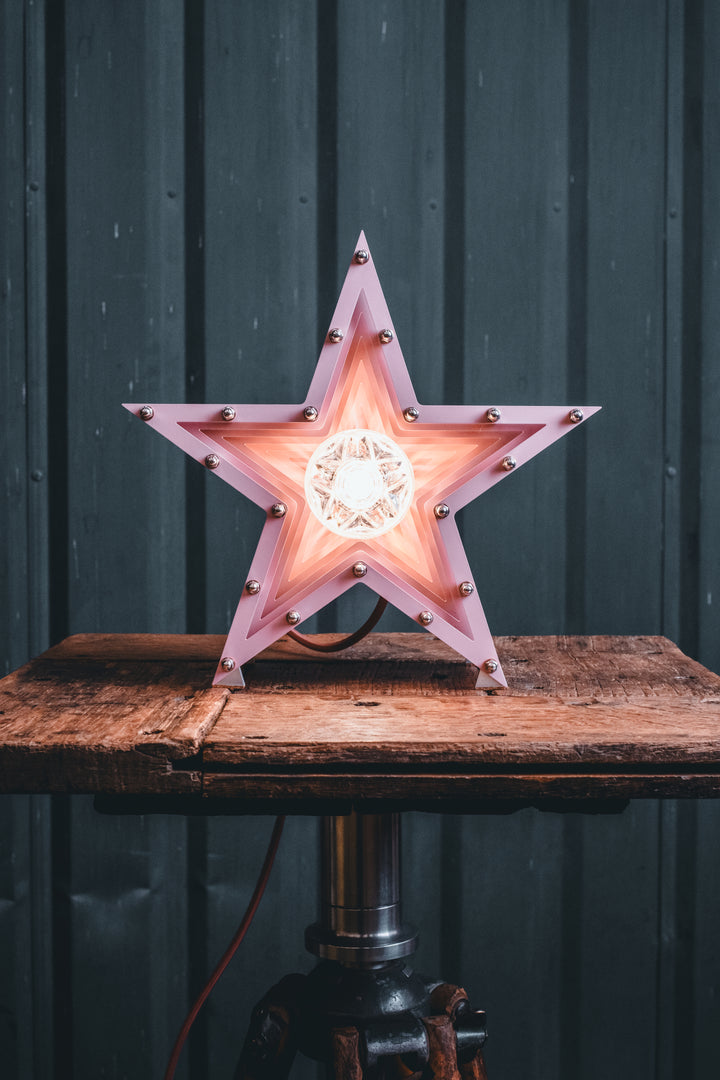 Handcrafted pink star-shaped lamp with layered metal fins and exposed central bulb, sitting on a rustic wooden table.