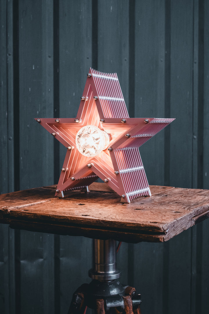Handcrafted pink star-shaped lamp with layered metal fins and exposed central bulb, sitting on a rustic wooden table.