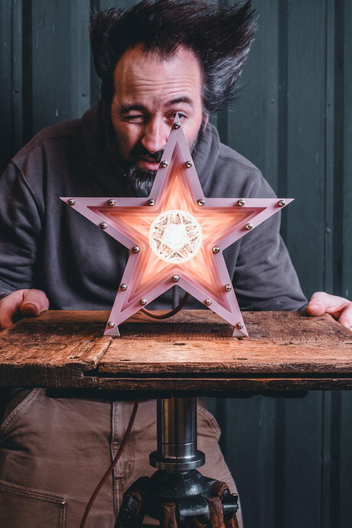 Handcrafted pink star-shaped lamp with layered metal fins and exposed central bulb, sitting on a rustic wooden table.